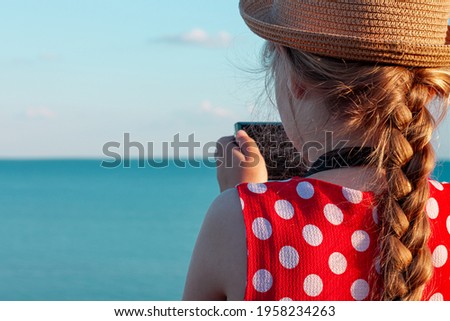 Little photographer child in straw hat and red polka-dot dress on vintage bench taking picture of soft pink rabbit toy on sea lanscape background. Girl looks at camera in hands. Friendship fun travel.