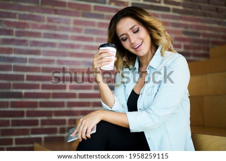 Cheerful hipster girl with takeaway caffeine beverage and modern cellphone gadget enjoying own web popularity resting outdoors, happy young female millennial with coffee to go and mobile phone