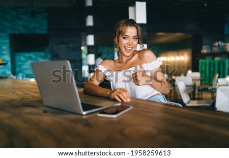 Optimistic smiling young lady in trendy white top sitting at high table with contemporary netbook and smartphone in restaurant while looking at camera and showing thumb up sign