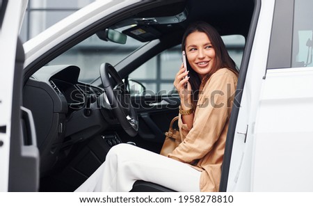 Talks by phone with door opened. Fashionable beautiful young woman and her modern automobile.
