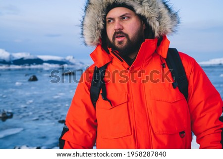 Adult bearded male in warm red jacket looking away while walking in solitude on coastline with frozen sea on background