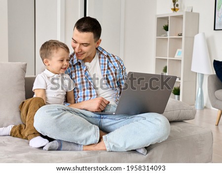 Happy young dad and cute small kid son using laptop computer at home. Smiling father distance learning or working online with toddler child, watching cartoons having fun spending time sitting on couch