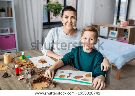 family, creativity and craft concept - mother and little son with glue sticks and paper making pictures of dry autumn leaves, pine cones and chestnuts at home