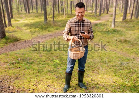 picking season and leisure people concept - happy smiling middle aged man with wicker basket and smartphone using app to identify mushroom in autumn forest