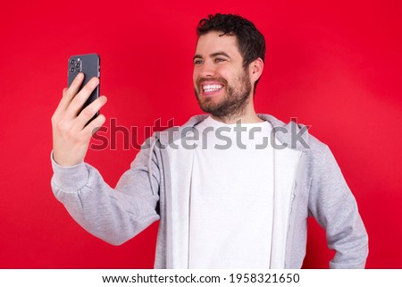 Isolated shot of pleased cheerful young handsome caucasian man in sports clothes against red background, makes selfie with mobile phone. People, technology and leisure concept
