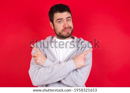 young handsome caucasian man in sports clothes against red background crosses arms and points at different sides hesitates between two items or variants. Needs help with decision