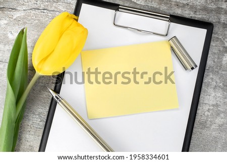 On a wooden table there is a stationery tablet with a yellow blank sheet near which there is a tulip. Postcard. Holiday background
