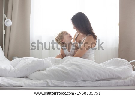 Beautiful blond child and his mother, playing at home in the morning in bed, smiling, laughing and having fun together