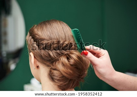 Professional hairdresser making coiffure for female client in front of big mirror. Work process in beauty studio. Bride getting ready for wedding in beauty salon. Close-up picture of fancy hairdo.
