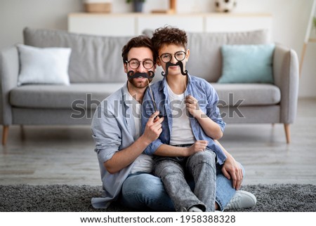 Father best example of success and courage. Son and dad holding fake moustache on sticks and smiling to camera, having fun at home, sitting on floor in living room Royalty-Free Stock Photo #1958388328