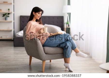 Free Time To Relax. Portrait of beautiful calm woman sitting on comfortable chair at home, reading paper book, holding cup and drinking hot coffee. Happy lady taking break from work, copy space Royalty-Free Stock Photo #1958388337
