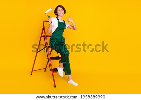 Full size photo of happy positive girl sit ladder hold roller showing thumb-up excellent isolated on yellow color background