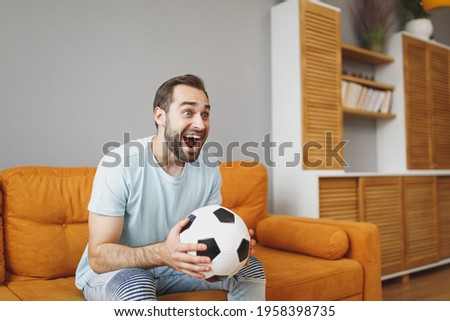 Excited young bearded man football fan 20s wearing basic blue t-shirt cheer up support favorite team with soccer ball keeping mouth open sitting on couch resting spending time in living room at home