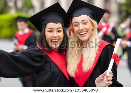 Two cheerful multiracial young ladies girlfriends in graduation clothes taking selfie, happy female students showing diplomas and smiling at camera, closeup portrait over university campus