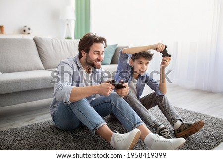 Videogames concept. Joyful father and son competing in online games, holding joysticks while sitting on carpet in living room and spending time together on weekend, copy space Royalty-Free Stock Photo #1958419399