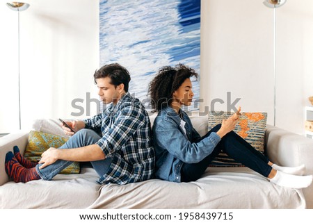 Disagreement between a loving multiracial couple. Guy and a girl offended at each other are sitting on the sofa with their backs to each other, using smartphones,dependent on Internet and social media Royalty-Free Stock Photo #1958439715