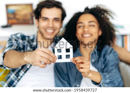 New home owners. Joyful multiethnic couple holding paper house, sitting on a sofa in their new modern apartment or house. Defocused african american girl and hispanic guy happy to buy their own home Royalty-Free Stock Photo #1958439727