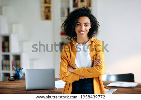 Portrait of a pretty African American girl. Confident stylish young african american woman with curly hair, stands near the desktop with crossed arms, looks at the camera, smiles friendly Royalty-Free Stock Photo #1958439772