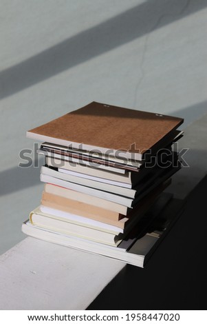 Stack of closed books lying on top of each other on a white stone concrete parapet. Gray white background. Black shadow on the right side of the photo.