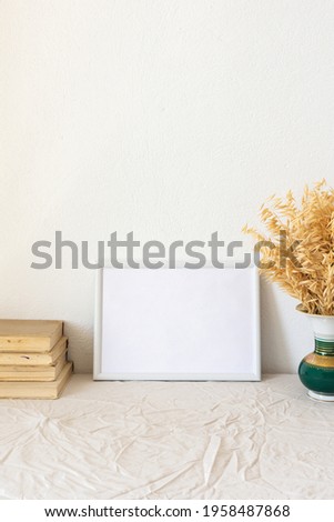 Home decor mock-up, blank picture frame near white painted concrete wall ,vase with dried yellow oat stalks, old books
