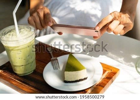 Woman hands using smartphone to take a picture of Matcha cheese cake with iced Matcha latte on white table in sunny day. 
