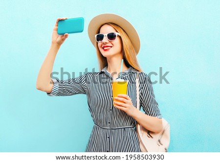 Portrait of smiling young woman with cup of juice taking a selfie by smartphone wearing a summer straw hat and backpack on a blue background