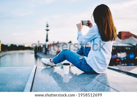 Young millennial woman using smartphone application for making photo while resting at urban setting, back view of female hipster blogger in casual wear shooting video on digital mobile phone