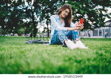 Caucasian hipster girl in classic eyewear using cellphone gadget for online messaging connect to 4g wireless in park, happy female blogger networking social media while installing smartphone app