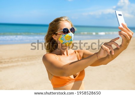 Funny girl taking selfie photo by smartphone on tropical sea beach. New rules to wear cloth face covering mask at public places due coronavirus COVID 19. Family holidays with children, summer travel.