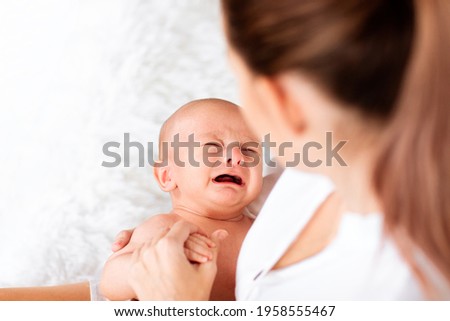 Young pretty mother with tail in white shirt holding crying newborn baby. Focus is at the baby. Copy space. Royalty-Free Stock Photo #1958555467
