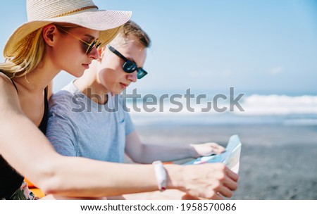 Side view of young female and male in sunglasses and casual wear looking at map on sea shore while spending time together