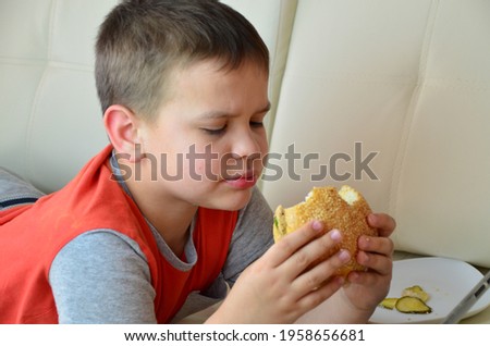 boy at home on the couch, eating a hamburger and looking at the tablet, watching cartoons or talking with friends he eat burgers and look at something on the tablet. he focused on screen