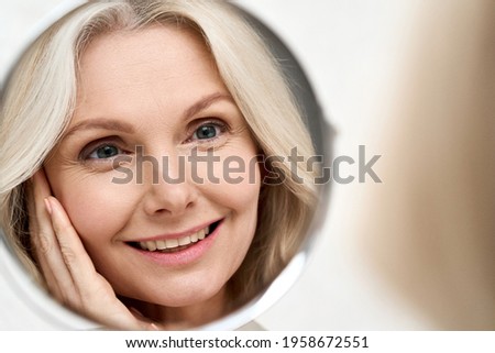 Happy middle 50 years aged woman with blond hair looking at mirror reflection examining and enjoying antiaging beauty treatments. Beauty hydrate skin care wrinkle prevention concept. Royalty-Free Stock Photo #1958672551