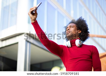 Young African American man taking a picture of himself with a smart phone. He is outdoors next to a modern building. Space for text. 