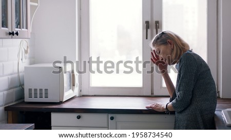 Weak lonely elderly woman leaning on the kitchen counter and looking through the window. High quality photo