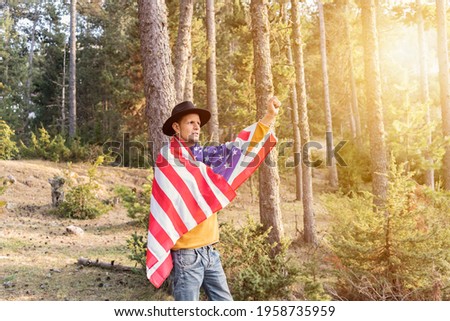 Young patriotic man in cowboy hat holding United States American flag and raising fist and arm in forest.Beautiful woodland landscape at sunset.Independence day celebration concept, 4th of July.