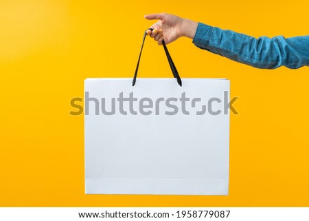 Close up cropped shot of female hand holding plain white shopping bag on bright colored yellow background. Mock up, copy space for your text or logo.