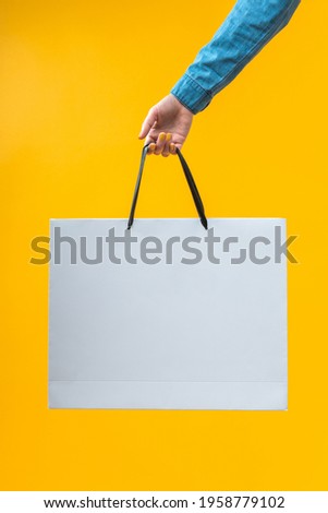 Vertical shot of female hand holding plain white shopping bag isolated on bright colored yellow background. Mock up, copy space for your text or logo.