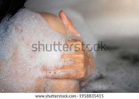 A woman is taking shower in bathtub, action during using hand with soap foam to cleaning back shoulder part, Relaxing and skin care concept. Close-up at people's hand and soft focus photo.