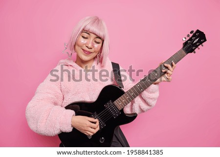 People hobby music concept. Pleased stylish pink haired talented female musician plays rock n roll song on acoustic guitar performs at stage being popular star isolated over rosy studio background