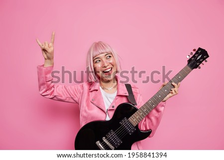 Energetic happy rock star keeps arm raised makes heavy metal sign happy to write own album with popular songs plays acoustic guitar dressed in jacket isolated over pink background. Cheerful frontwoman