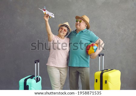 Happy old couple making dream come true. Smiling senior tourists in sunglasses and sun hats holding paper passenger airplane standing on grey studio background. Air flight and sea beach holiday Royalty-Free Stock Photo #1958871535
