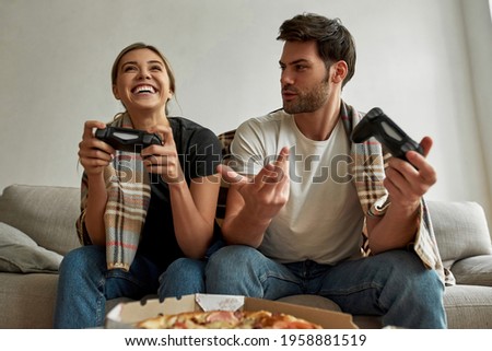 Picture of a young couple on self isolation having a console competition and eating pizza, both spending their time on self isolation, woman is looking happy cause winning, man has no idea how