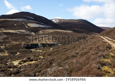 A landscape in the Scottish Highlands of 2 brown heather covered hills rising to a blue sky.  A track to the right of picture leads around a corner and further up the Scottish Glen.
