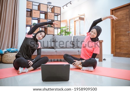 two women wearing hijab sportswear sit cross-legged on the floor with their bodies leaning to the side and hands up while warming up their arms together in the house