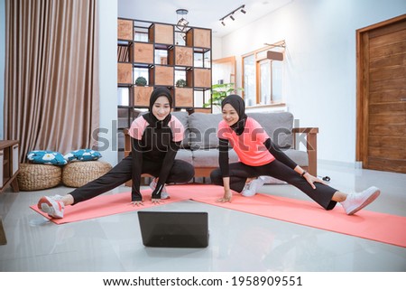 two young woman wearing a hijab gym clothes squatting stretches with one leg pulled sideways while in front of a laptop in the house