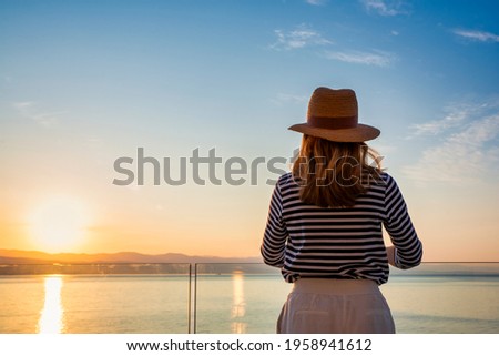 Shot of pretty woman wearing striped sweater and straw hat while standing on balcony and looking at sea view. Daydreaming.