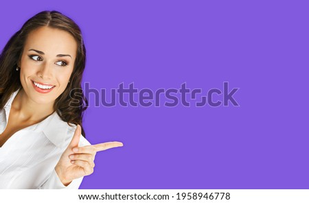 Young happy smiling woman in white confident blouse, showing pointing advertising, copy space for some text. Business ad concept. Violet purple background. Brunette businesswoman. 