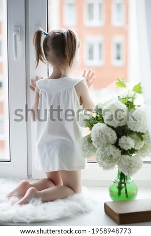 A little girl is sitting on the windowsill. A bouquet of flowers in a vase by the window and a girl sniffing flowers. A little princess in a white dress with a bouquet of white