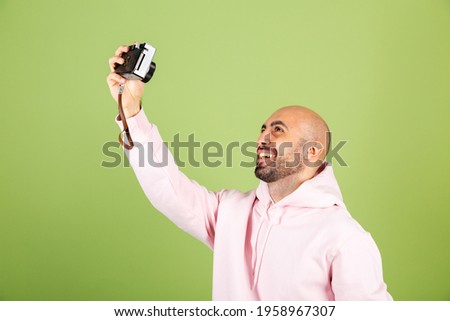 Young bald caucasian man in pink hoodie isolated on green background positive cheerful take photo selfie on professional camera
